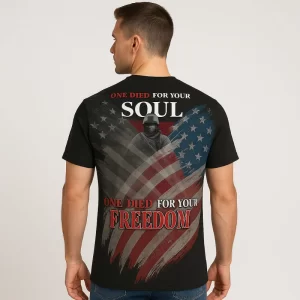 A black short-sleeve t-shirt with an all-over American flag graphic in muted red, white, and blue tones. On the back, a soldier silhouette overlays the flag with the large bold text: 'One Died For Your Soul, One Died For Your Freedom.' The design is strong and patriotic, blending faith and military honor.